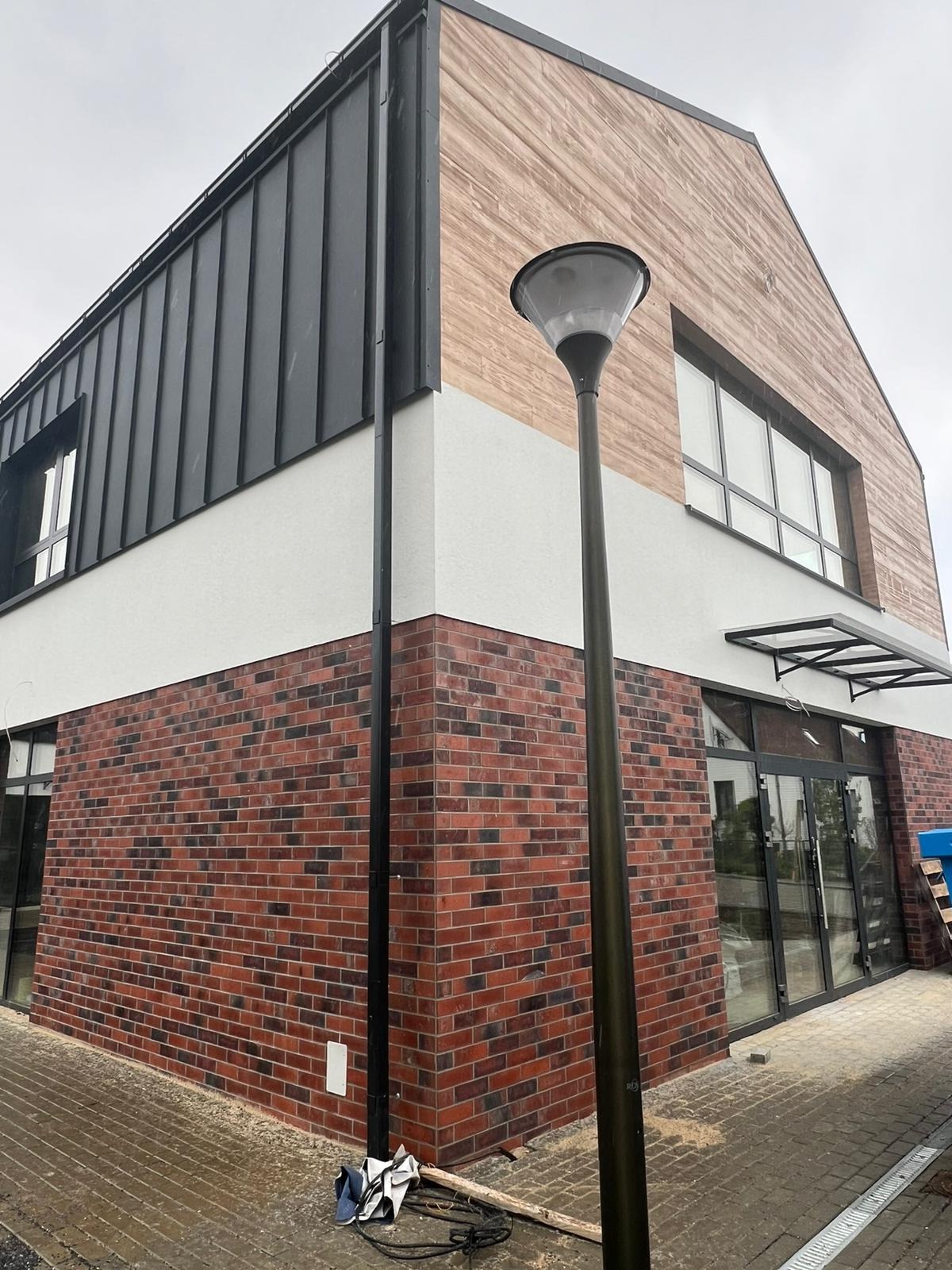 Nowoczesna elewacja budynku z cegły klinkierowej, tynku i paneli drewnopodobnych. Widoczny fragment chodnika i lampa uliczna. Ujęcie z dołu, pochmurny dzień.