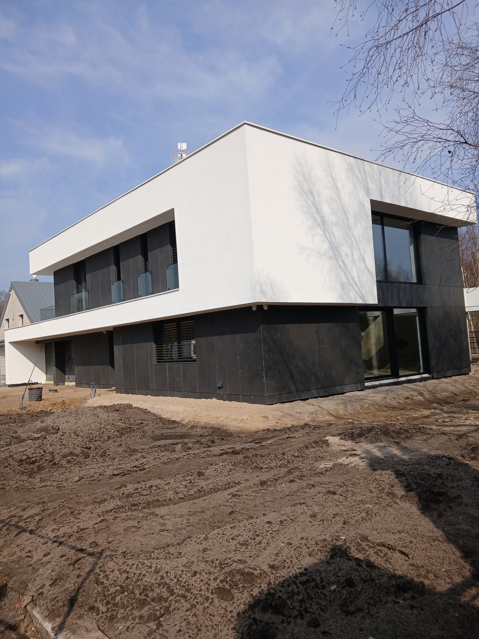 Nowoczesny dom z biało-czarną elewacją, duże okna, minimalistyczny design. Widoczne ślady prac ziemnych wokół budynku. Antena na dachu.