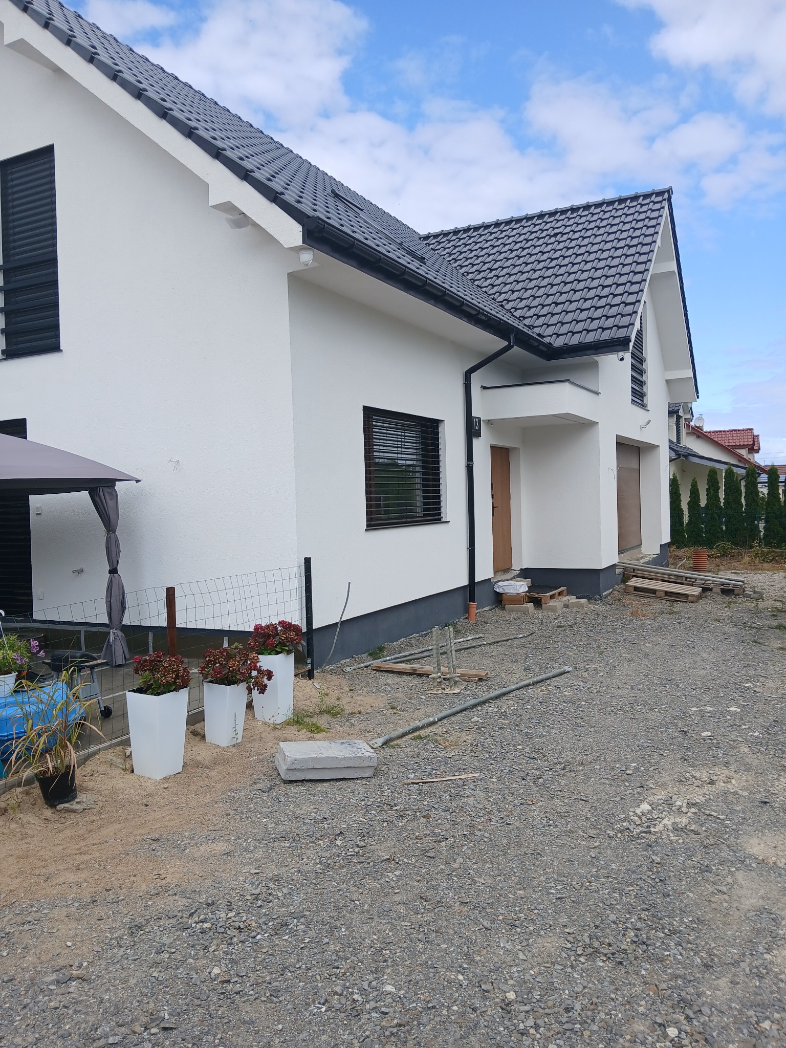 Biały dom jednorodzinny z ciemną dachówką i szarą podmurówką, widok z boku, z elementami wykończeniowymi i roślinami w donicach na pierwszym planie.