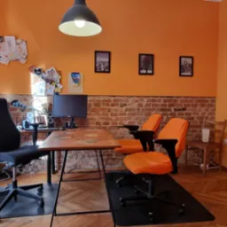 Pomarańczowy pokój z biurkiem ozdobionym naklejkami, monitor, krzesła biurowe i ściana z cegły. Na ścianie rysunki dziecięce i zdjęcia.