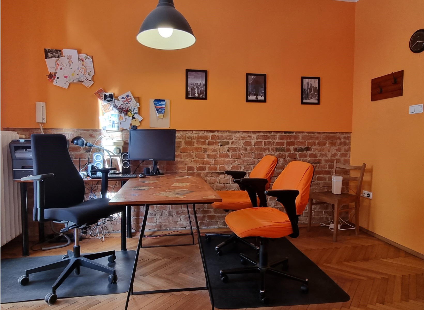 Pomarańczowy pokój z biurkiem ozdobionym naklejkami, monitor, krzesła biurowe i ściana z cegły. Na ścianie rysunki dziecięce i zdjęcia.