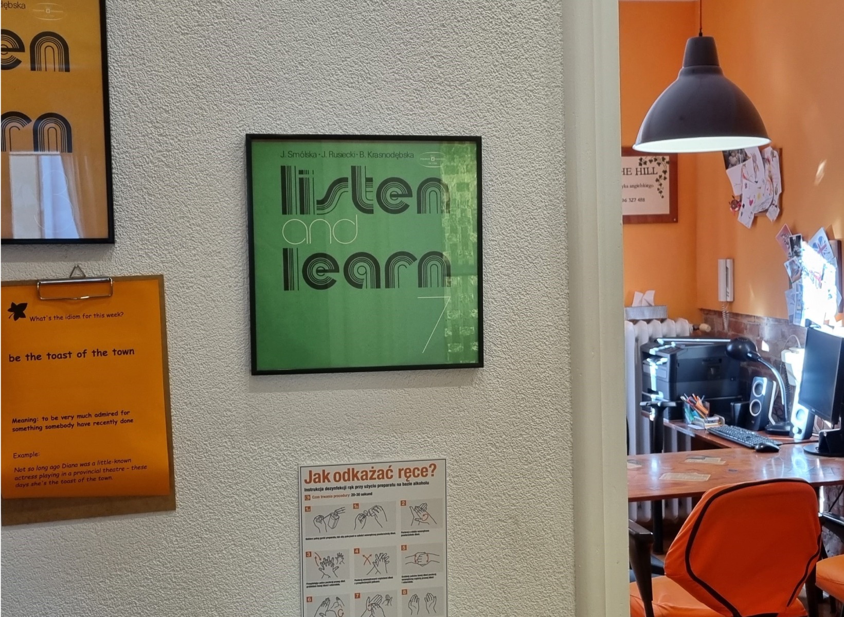 Sala językowa: plakat 'Listen and Learn', tablica z idiomem 'be the toast of the town', instrukcja dezynfekcji rąk. Widok na biurko i pomarańczowe krzesło.