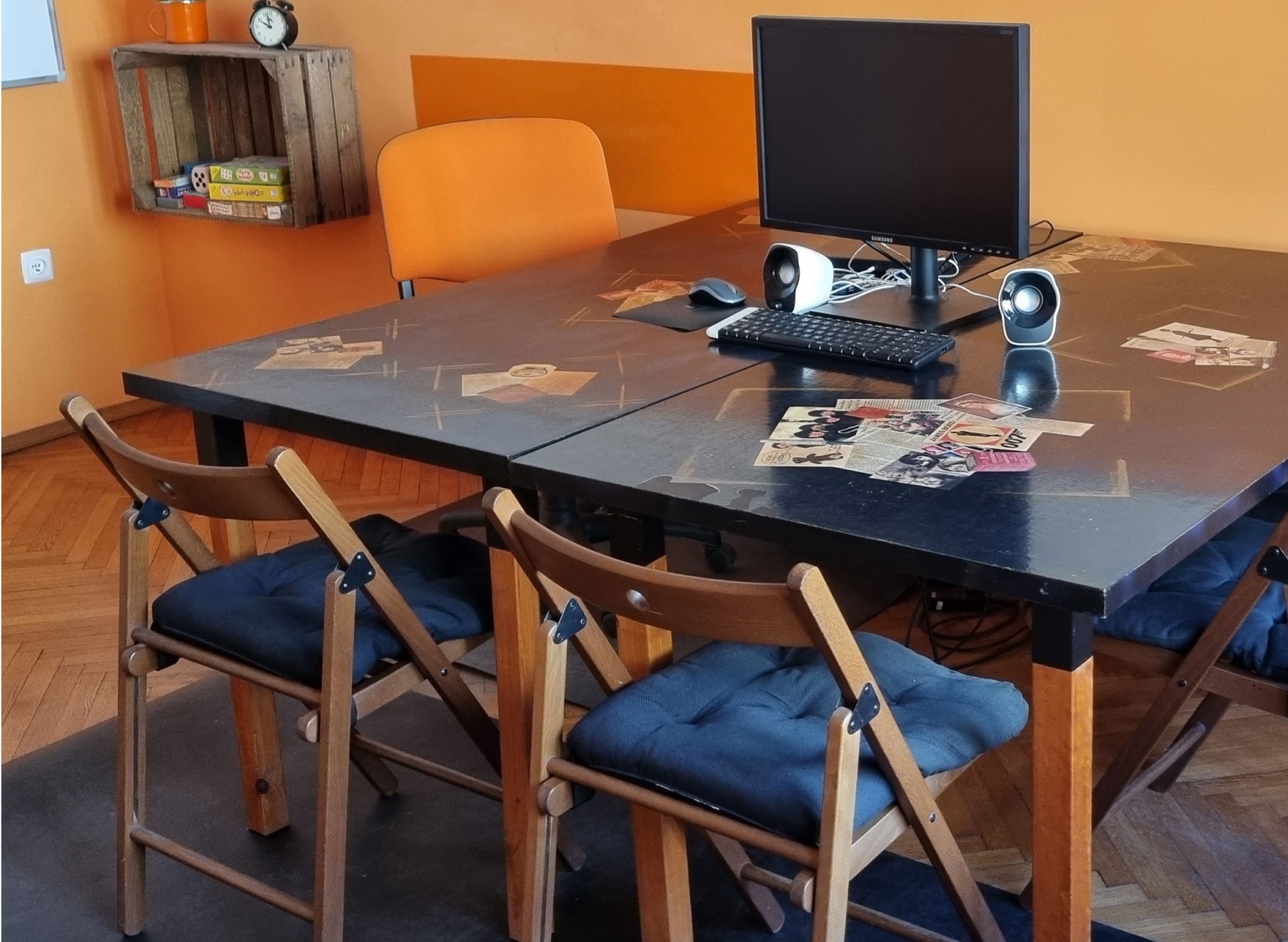 Stół z komputerem i krzesłami w pomarańczowym pokoju, potencjalne miejsce nauki angielskiego. Dekorowany blat, pudełko na ścianie, przytulna atmosfera.