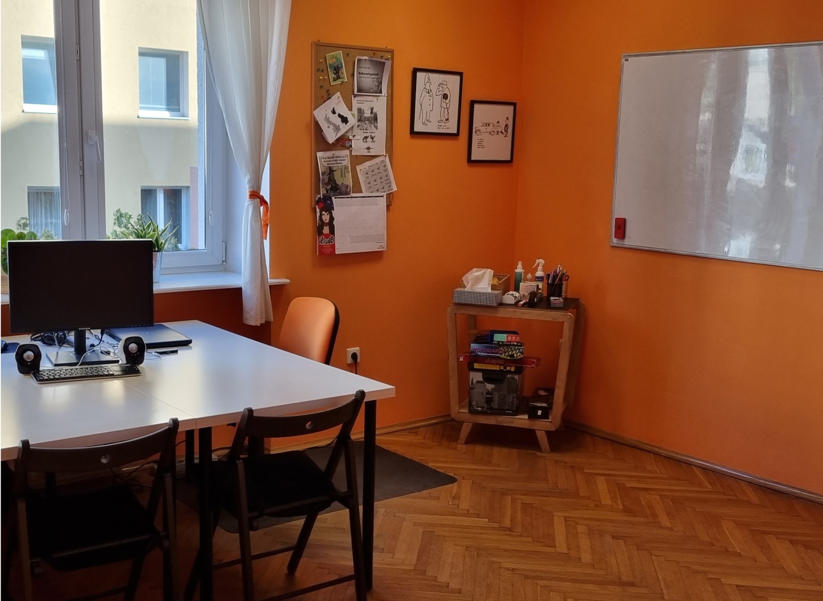 Sala lekcyjna z pomarańczowymi ścianami, białym stołem, krzesłami i tablicą. Na ścianie korkowa tablica z notatkami i kalendarzem.