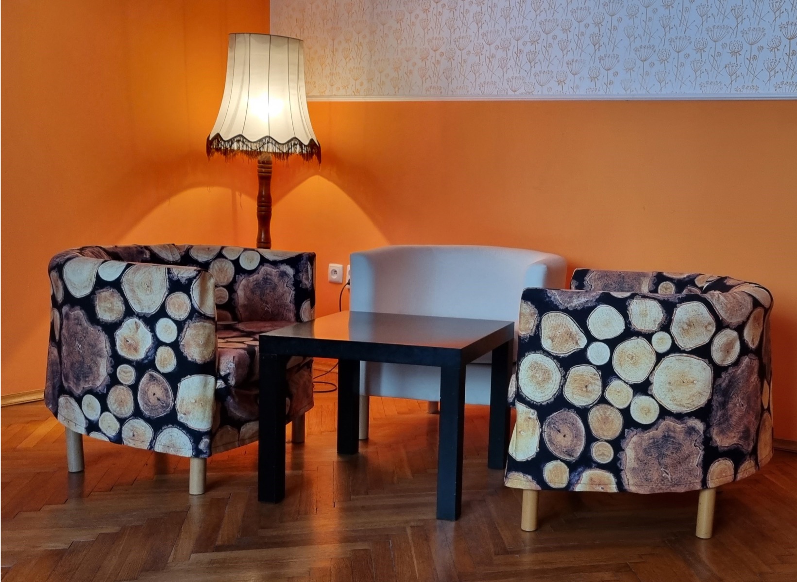 Pomarańczowe wnętrze z lampą, dwoma fotelami w wzór drewna i czarnym stolikiem. Biała sofa w tle. Przytulna aranżacja pokoju.