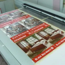 Arkusz z powtarzającymi się grafikami mebli i pomieszczeń (sypialnia, jadalnia, salon) z logo Black Red White, umieszczony na stole plotera UV do druku wielkoformatowego.