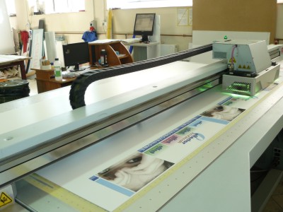 Proces druku UV na płaskim podłożu za pomocą przemysłowej drukarki wielkoformatowej, widoczne detale drukowanego materiału z grafiką.