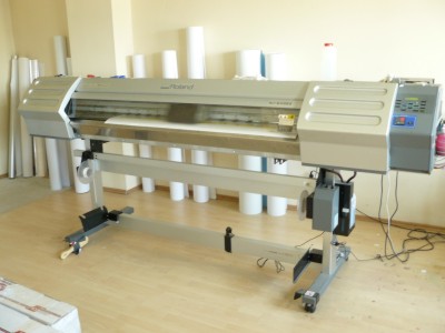Szerokoformatowa drukarka Roland z białym papierem w trakcie druku, widoczne rolki z materiałem do druku w tle.