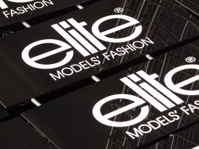 Czarne, błyszczące naklejki z białym logo 'Elite Models' Fashion' ułożone w rzędzie, widoczne odbicia światła.