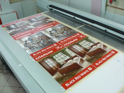 Arkusz z powtarzającymi się grafikami mebli i pomieszczeń (sypialnia, jadalnia, salon) z logo Black Red White, umieszczony na stole plotera UV do druku wielkoformatowego.