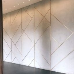 Epoxon - Ściana z mikrocementu w geometryczne wzory z metalicznymi listwami, oświetlona punktowo, z widoczną częścią ciemnej podłogi i zasłony w rogu.