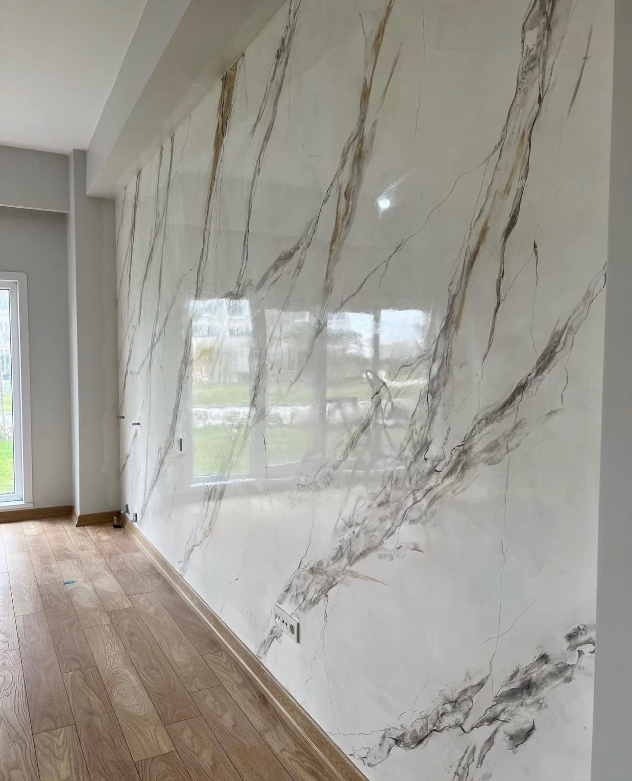 Ściana w salonie wykończona gładzią imitującą marmur z widocznymi żyłami w odcieniach szarości i złota, drewniana podłoga, okno w tle.
