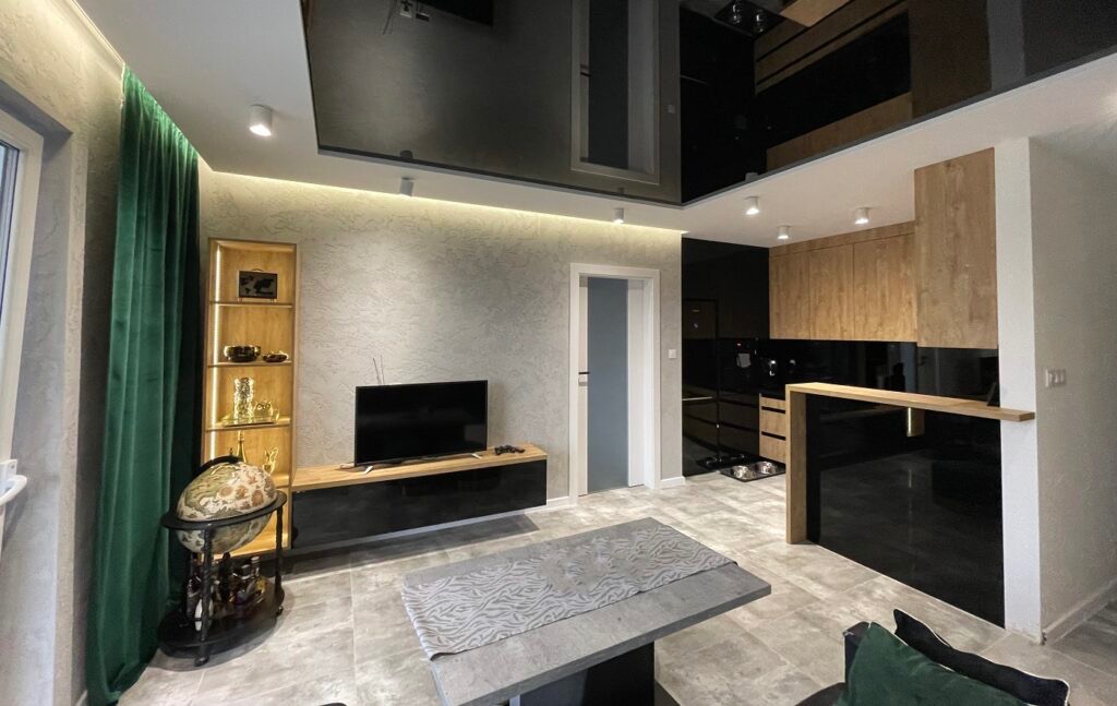 Eleganckie wykończenie salonu w Warszawie: szara ściana z fakturą, błyszczący sufit podwieszany i drewniane elementy zabudowy kuchennej. Nowoczesny design.