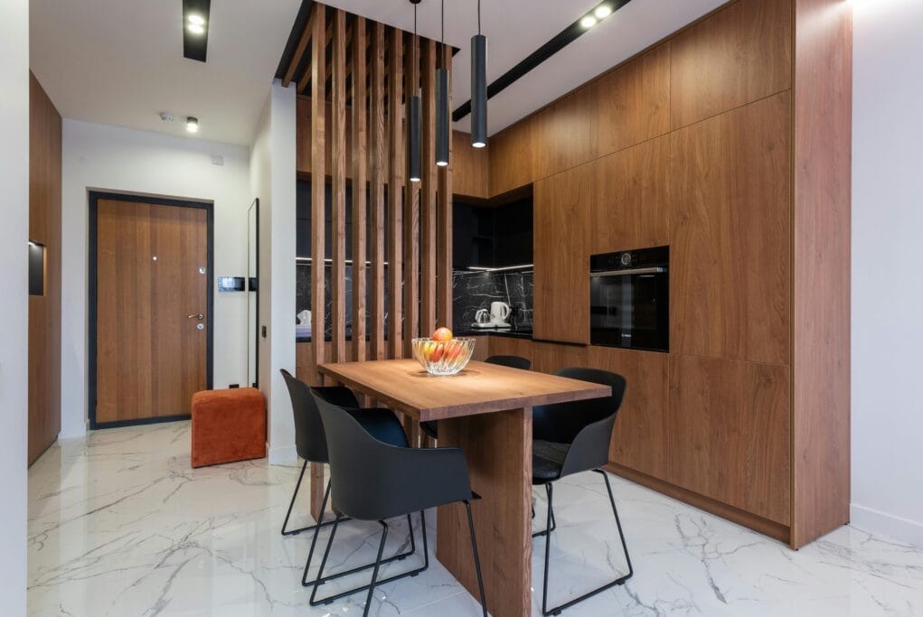 Nowoczesna kuchnia z drewnianymi szafkami, marmurową podłogą i stołem oddzielonym ażurową ścianką. Minimalistyczne lampy wiszące. Wykończenie wnętrza.