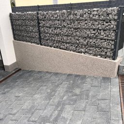 Budolix Paweł Wołoszyn - Gabion z kamieniami na posesji, z szarą elewacją i kostką brukową. Widoczne odpływy liniowe. Całość utrzymana w stonowanej kolorystyce.