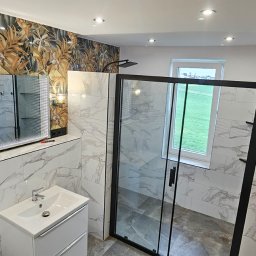 Budolix Paweł Wołoszyn - Nowoczesna łazienka z kabiną prysznicową typu walk-in, umywalką z szafką, lustrem z podświetleniem LED i grzejnikiem. Ściany wyłożone płytkami imitującymi marmur.