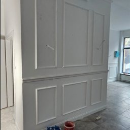 Budolix Paweł Wołoszyn - Eleganckie wykończenie ściany z dekoracyjnymi panelami w kolorze białym. Widoczne narzędzia malarskie. Szare płytki podłogowe. Wnętrze w trakcie prac.
