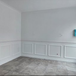 Budolix Paweł Wołoszyn - Eleganckie wykończenie wnętrza z białą sztukaterią na ścianach i szarą podłogą. Widoczne elementy instalacji elektrycznej i okno zabezpieczone folią malarską.