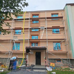 Budolix Paweł Wołoszyn - Ocieplanie budynku z rusztowaniem. Okna zabezpieczone folią. Widoczne wiadra z farbą i pracownicy przygotowujący się do malowania elewacji.