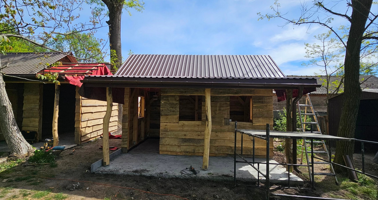 Budowa drewnianej konstrukcji z metalowym dachem, widoczne narzędzia i drabina. Konstrukcja w surowym stanie, otoczona zielenią.