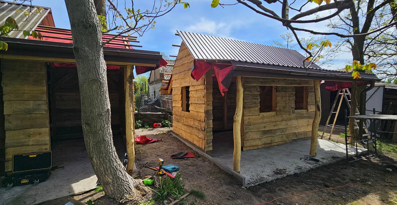 Budowa drewnianej konstrukcji z metalowym dachem, widoczne narzędzia i drabina, w tle budynki mieszkalne. Prace budowlane w toku, surowe wykończenie.