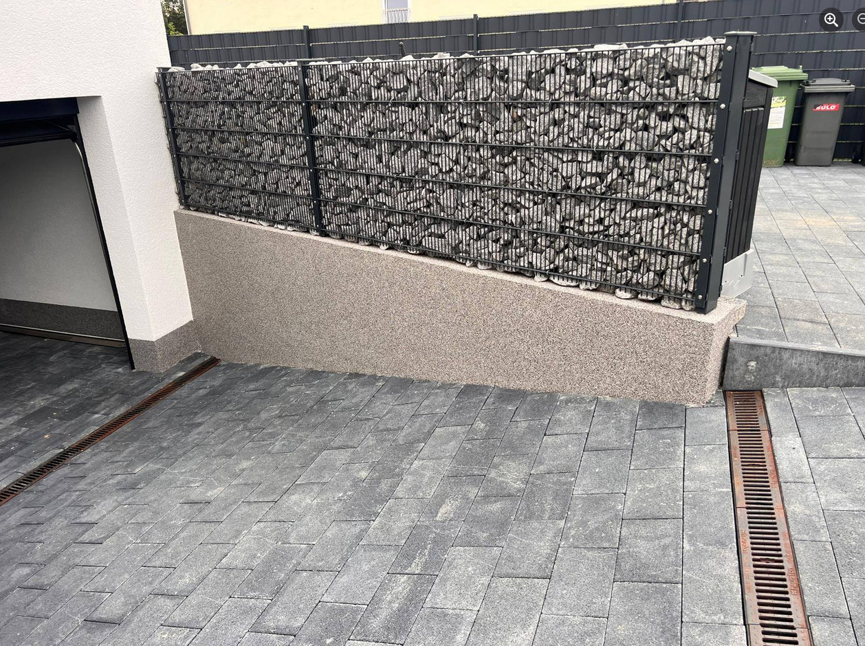 Gabion z kamieniami na posesji, z szarą elewacją i kostką brukową. Widoczne odpływy liniowe. Całość utrzymana w stonowanej kolorystyce.