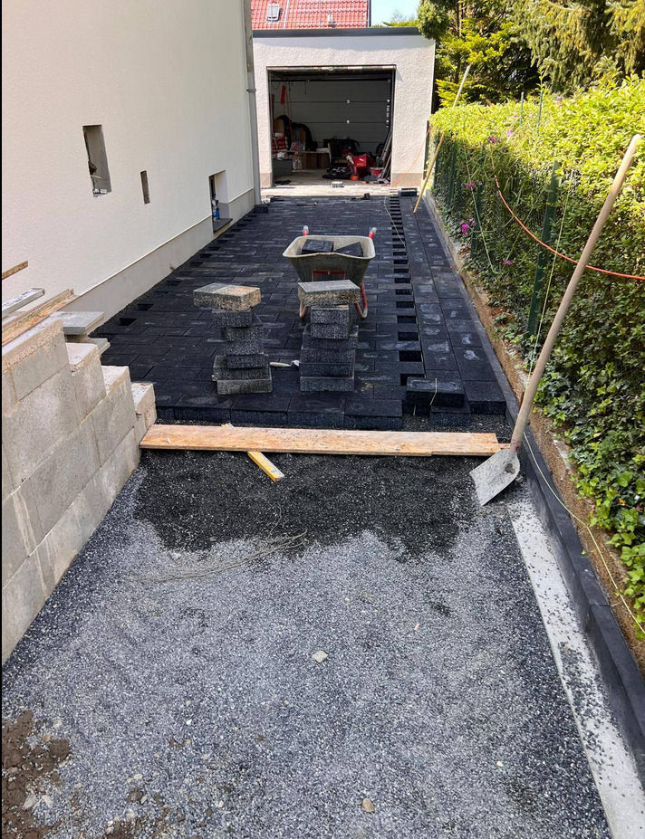 Układanie czarnej kostki brukowej na podwórku, widoczna taczka z kostką, częściowo ułożona nawierzchnia, przygotowane podłoże z grysu. Prace brukarskie w toku.