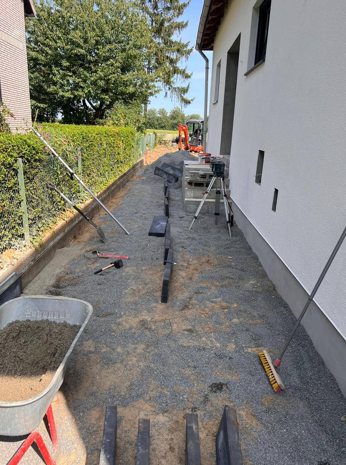 Układanie kostki brukowej na posesji: widoczny wózek z zaprawą, poziomica laserowa, ułożone obrzeża i częściowo kostka. W tle minikoparka.