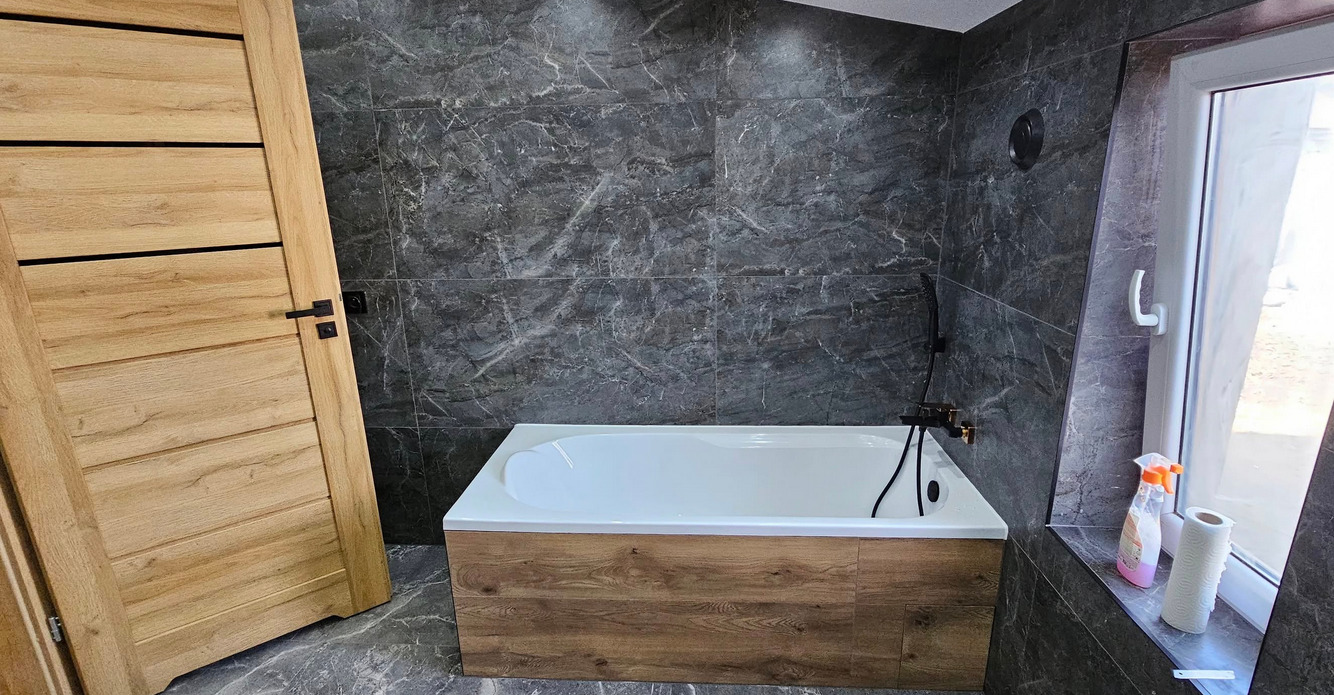 Nowoczesna łazienka z wanną obudowaną drewnem, szare marmurowe płytki, drewniane drzwi z czarnymi akcentami i oświetlenie punktowe.
