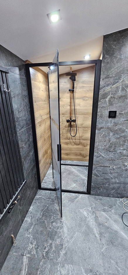 Nowoczesna kabina prysznicowa z czarną ramą i deszczownicą w odcieniach miedzi, wykończona płytkami imitującymi marmur i drewno. Otwarta szklana ścianka.