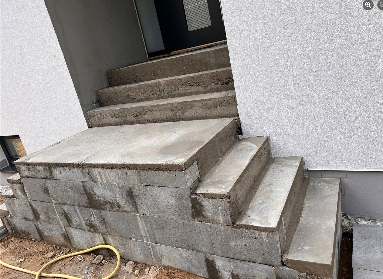 Betonowe schody wejściowe do budynku w trakcie budowy, z widocznymi bloczkami fundamentowymi i świeżym betonem. Surowy stan wykończenia.