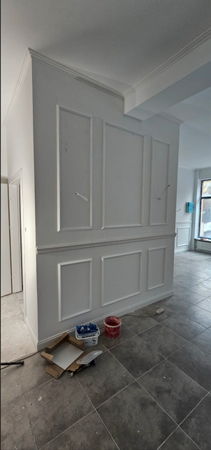 Eleganckie wykończenie ściany z dekoracyjnymi panelami w kolorze białym. Widoczne narzędzia malarskie. Szare płytki podłogowe. Wnętrze w trakcie prac.