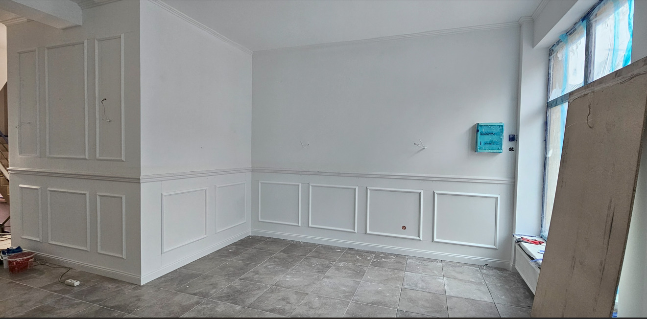 Eleganckie wykończenie wnętrza z białą sztukaterią na ścianach i szarą podłogą. Widoczne elementy instalacji elektrycznej i okno zabezpieczone folią malarską.