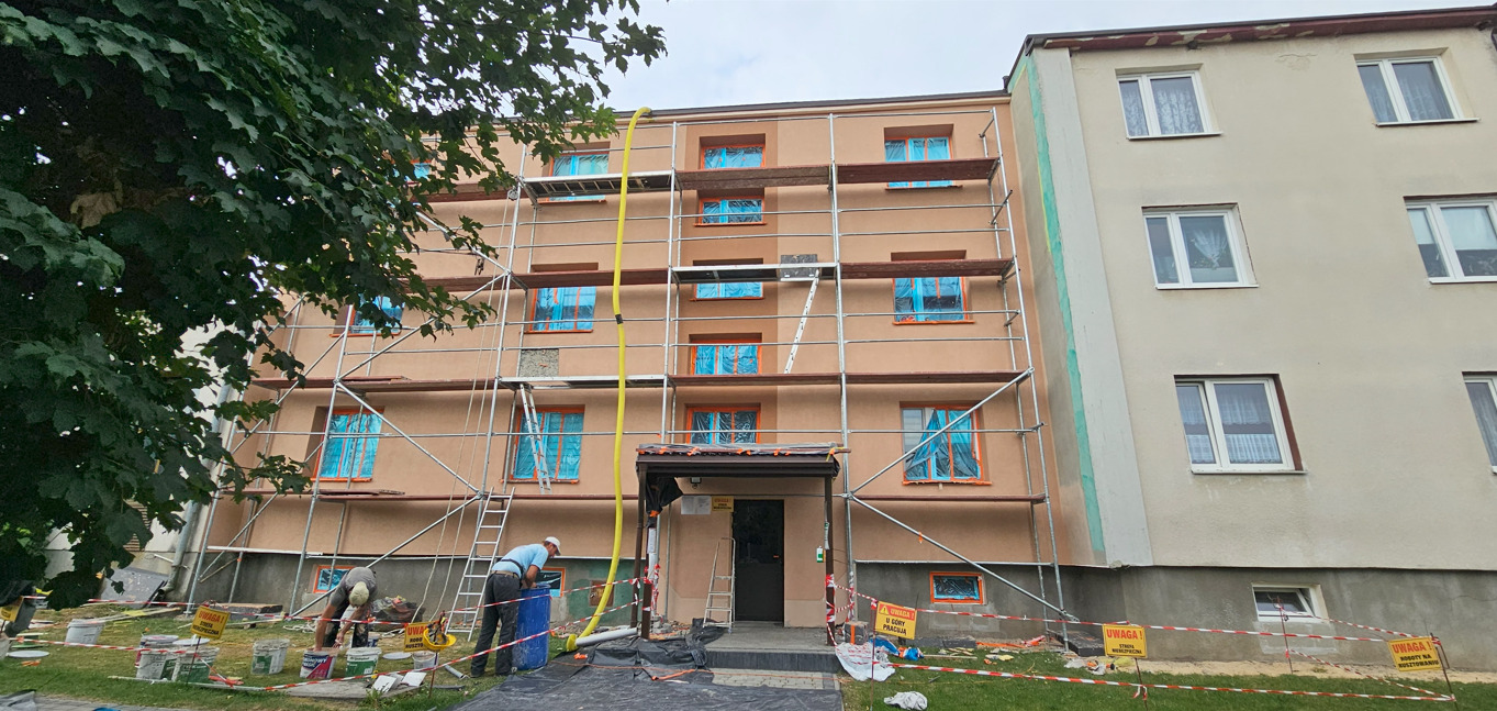 Ocieplanie budynku z rusztowaniem. Okna zabezpieczone folią. Widoczne wiadra z farbą i pracownicy przygotowujący się do malowania elewacji.