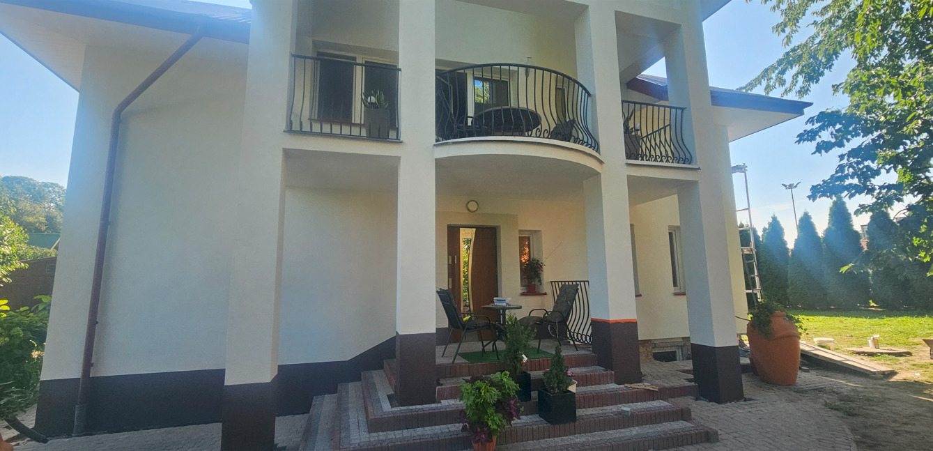 Odnowiona fasada domu z balkonem i kolumnami, widoczne detale wykończenia, schody wejściowe i elementy dekoracyjne. Jasna kolorystyka elewacji.