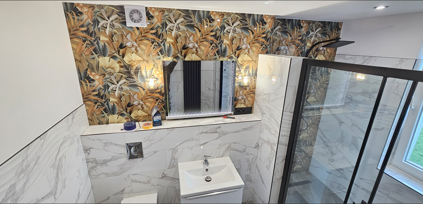 Wykończona łazienka z kabiną prysznicową typu walk-in, umywalką i lustrem z podświetleniem. Na ścianach płytki imitujące marmur i tapeta z motywem roślinnym.