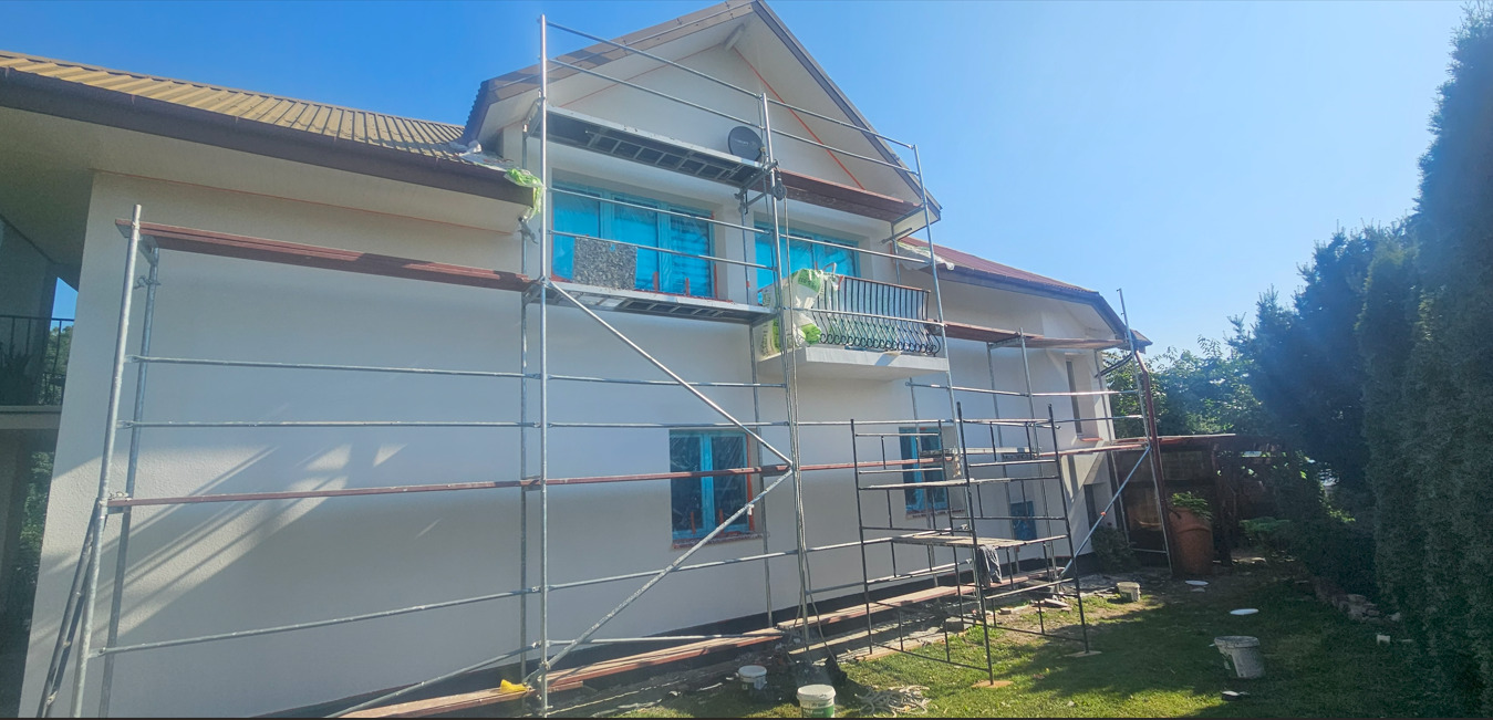 Elewacja domu jednorodzinnego w trakcie prac wykończeniowych, z rusztowaniem, zabezpieczonymi oknami i widocznymi śladami farby. Słoneczny dzień.