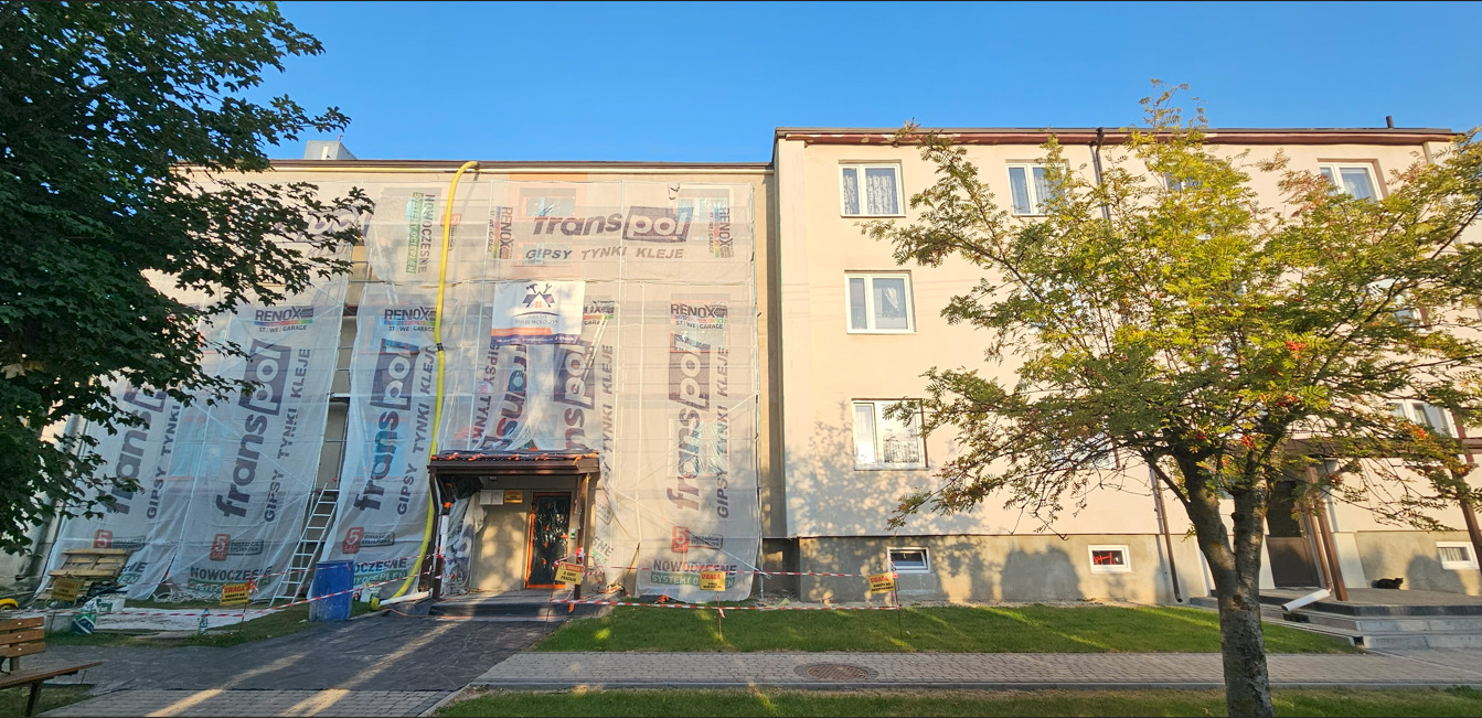 Budynek w trakcie prac remontowych, osłonięty siatką z reklamami. Widoczne rusztowania, drzewa i fragment chodnika. Ujęcie z dołu, w słoneczny dzień.