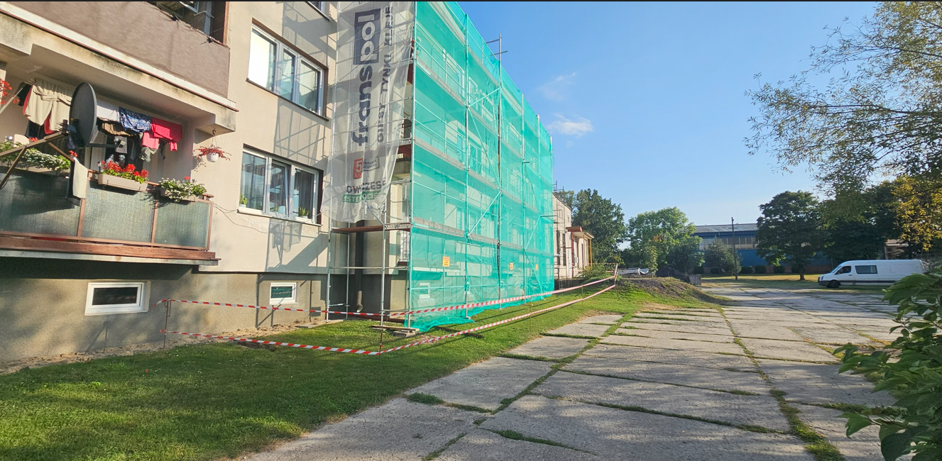 Blok w rusztowaniach osłoniętych siatką ochronną, widok z zewnątrz. Na balkonie suszarka z praniem. Taśma ostrzegawcza. Prace remontowe elewacji w toku.