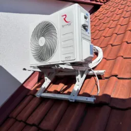 Biała jednostka zewnętrzna klimatyzacji Rotenso zamontowana na dachu z czerwonej dachówki, widok z dołu na tle błękitnego nieba.