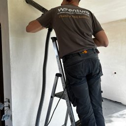 Wenturo Sp. z o. o. - Pracownik wierci otwór w ścianie pod instalację wentylacyjną. Widoczna drabina, przewód wentylacyjny i logo firmy na koszulce. Praca w trakcie realizacji.