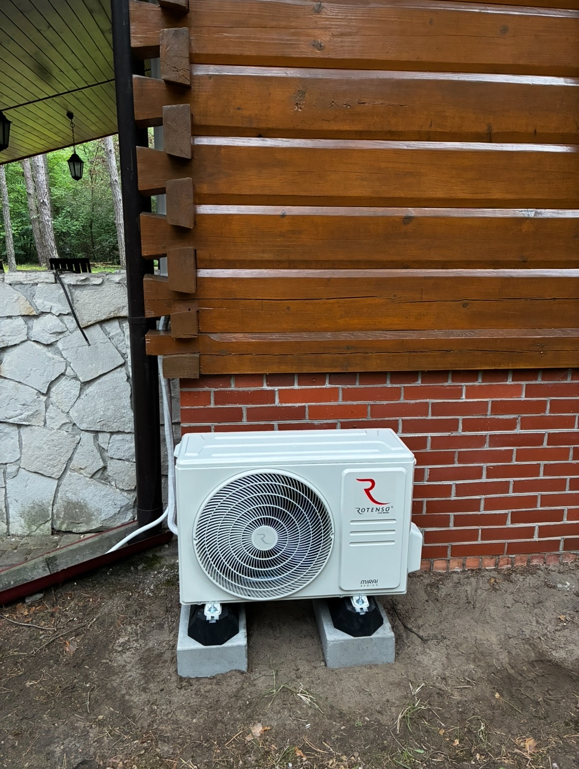 Biała jednostka zewnętrzna klimatyzacji Rotenso zamontowana na betonowych podstawach przy drewniano-ceglanej ścianie domu, z widocznym fragmentem kamiennej elewacji.