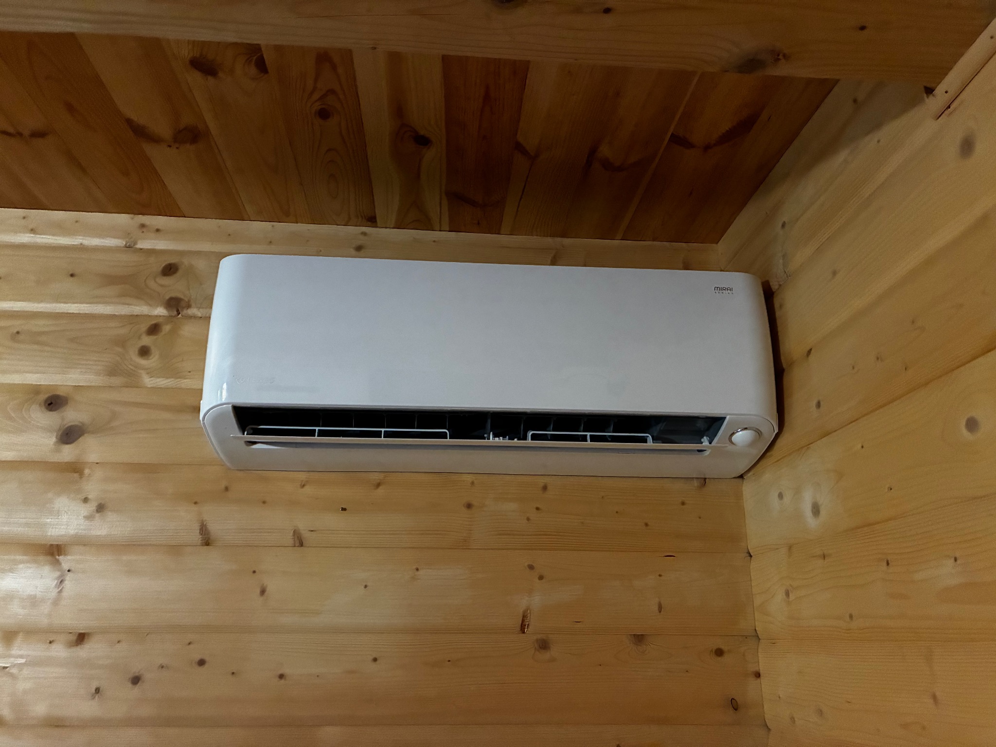 Biała klimatyzacja ścienna zamontowana w drewnianym wnętrzu. Widoczny fragment sufitu z boazerii. Kątowe ujęcie z dołu.