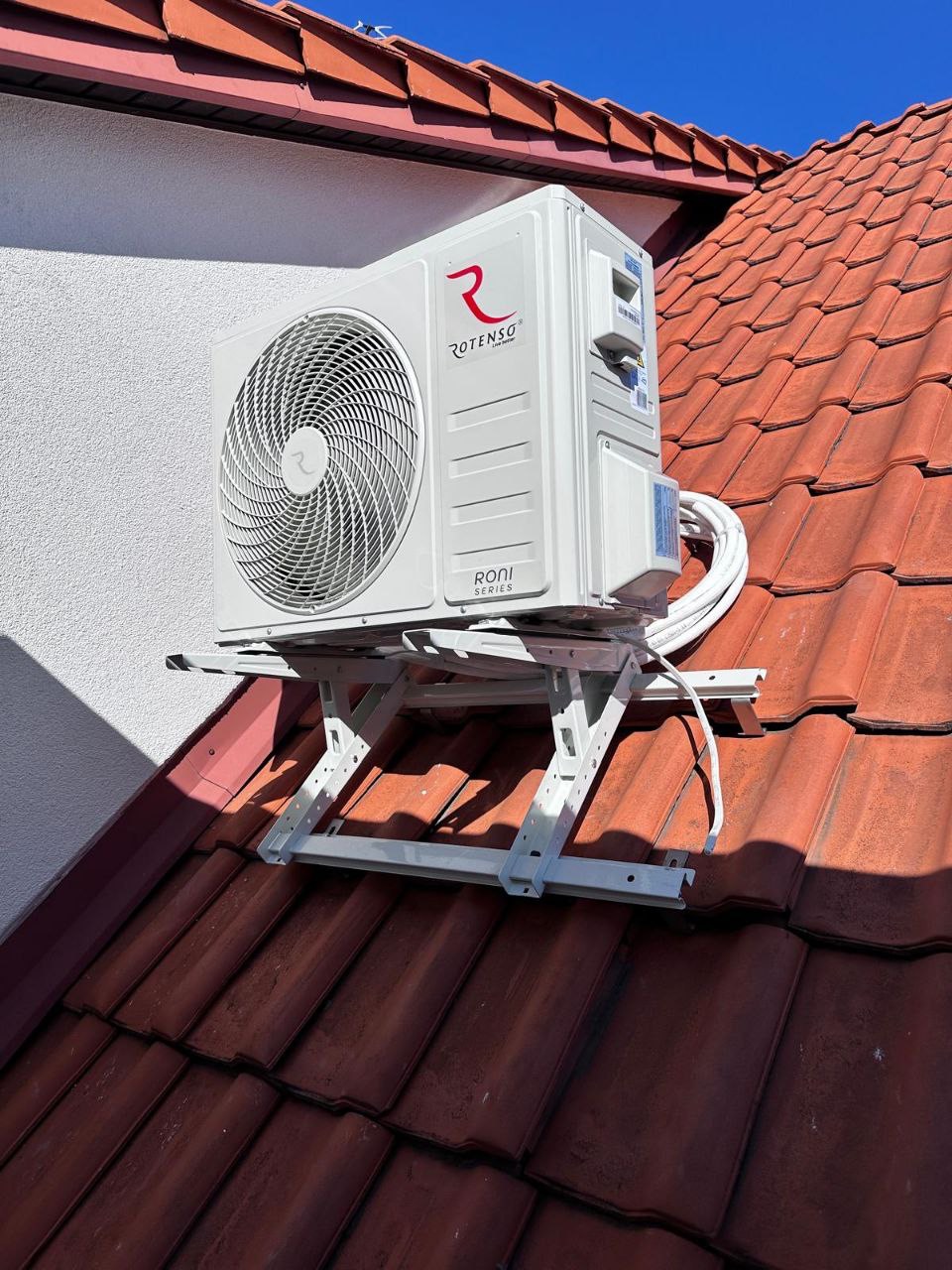 Biała jednostka zewnętrzna klimatyzacji Rotenso zamontowana na dachu z czerwonej dachówki, widok z dołu na tle błękitnego nieba.