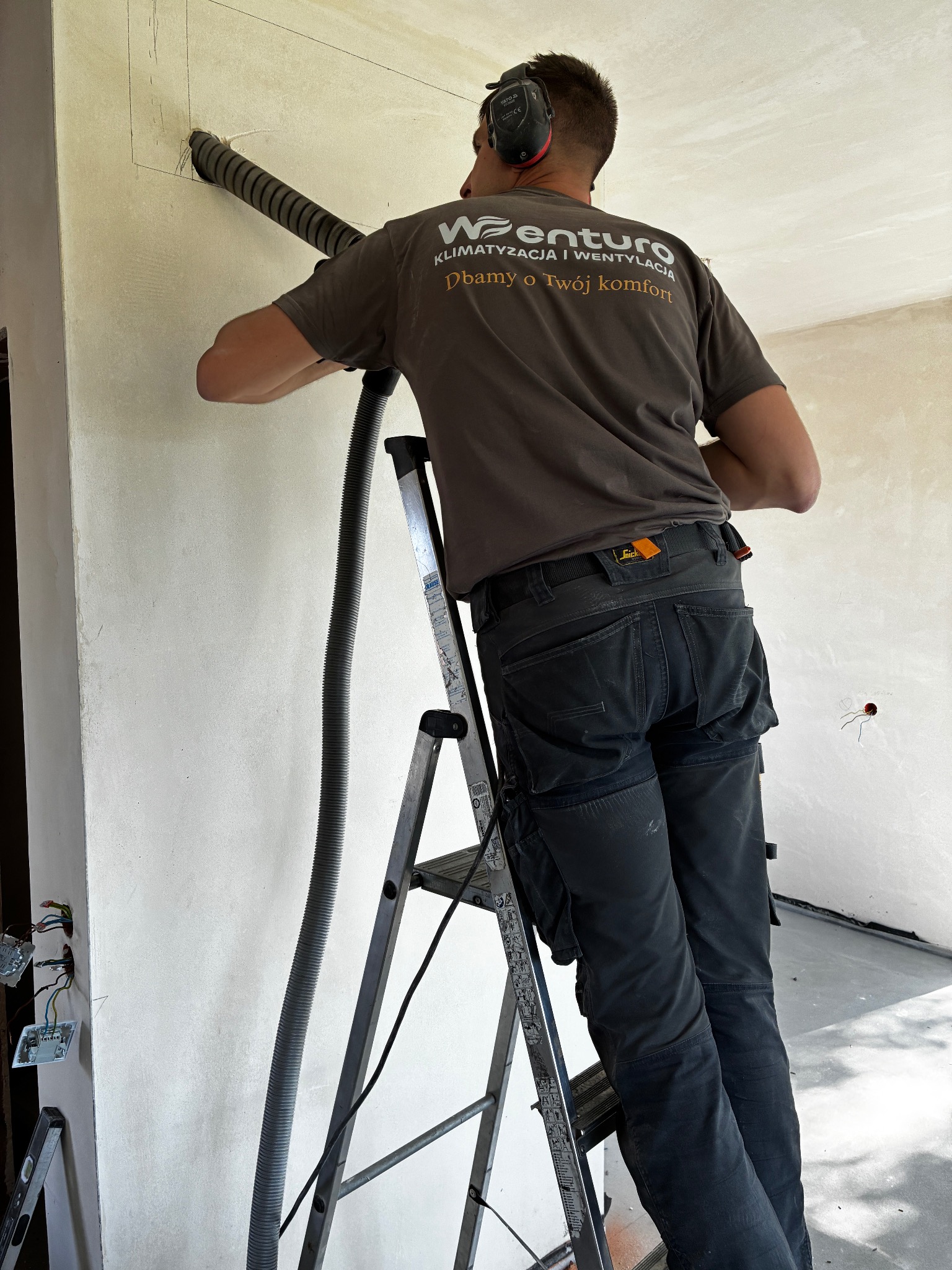 Pracownik wierci otwór w ścianie pod instalację wentylacyjną. Widoczna drabina, przewód wentylacyjny i logo firmy na koszulce. Praca w trakcie realizacji.