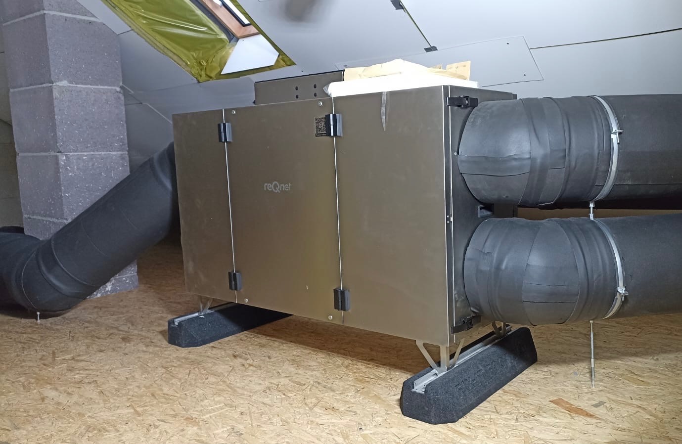 Centrala rekuperacyjna z podłączonymi przewodami wentylacyjnymi, zainstalowana na poddaszu z drewnianą podłogą i widocznym oknem dachowym.