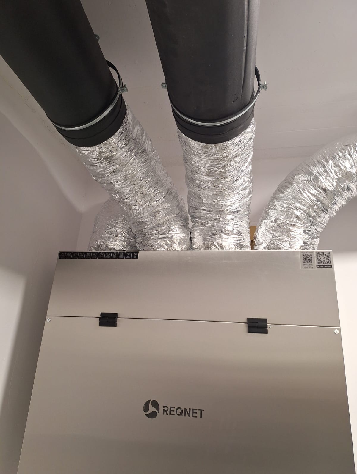 Centrala rekuperacyjna REQNET z podłączonymi przewodami wentylacyjnymi w pomieszczeniu o białych ścianach i suficie. Widok od dołu, centralnie.