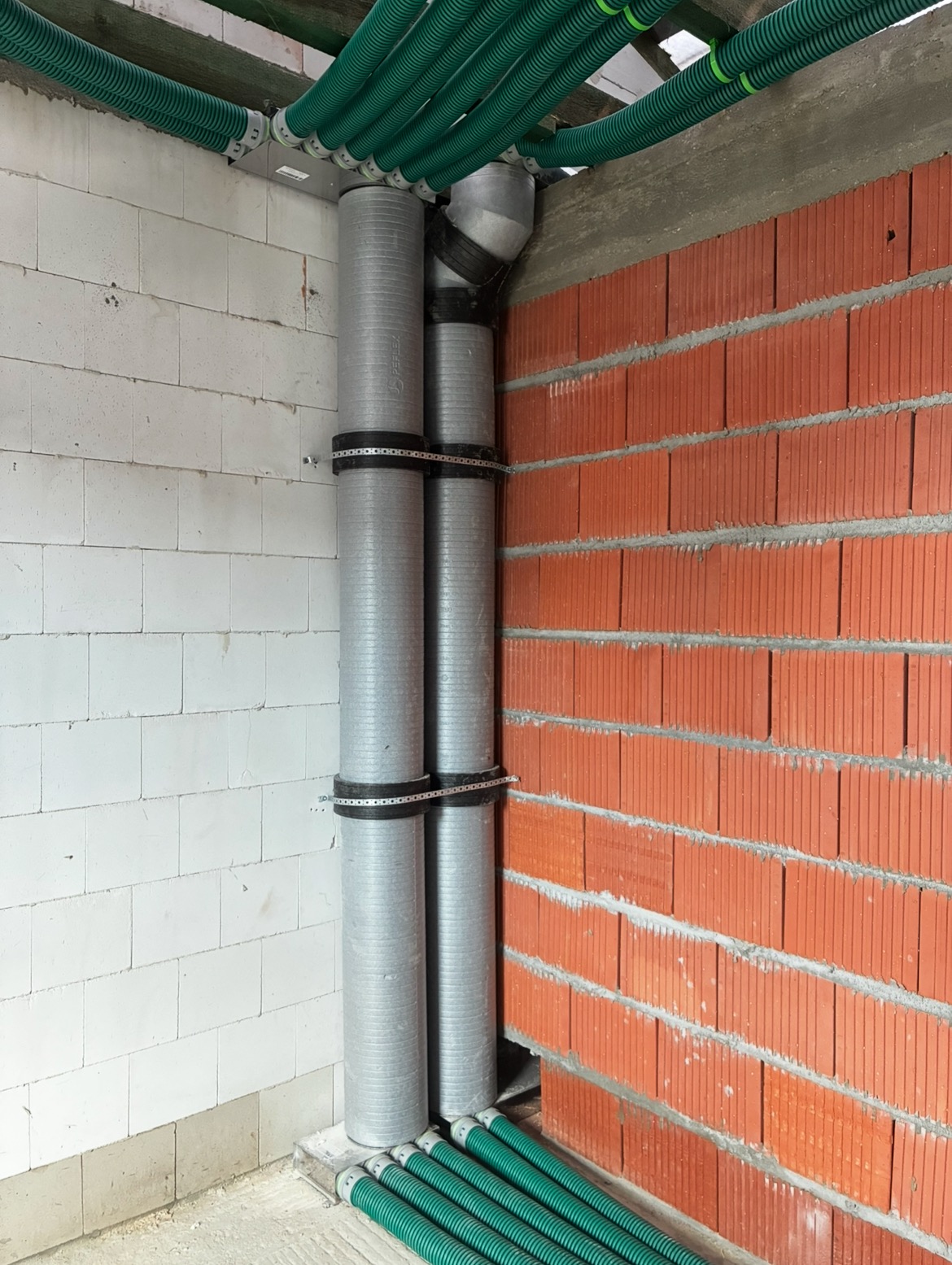 Montaż instalacji rekuperacji: zielone rury wentylacyjne pod sufitem i szare rury pionowe, mocowane do ściany z cegły i bloczków. Etap instalacji w surowym budynku.