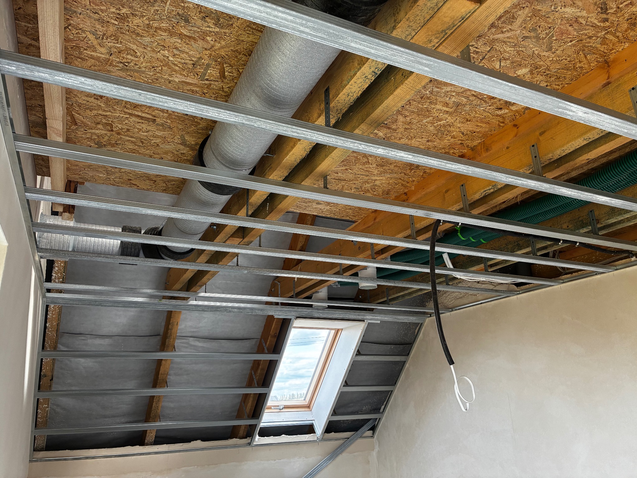 Szara rura wentylacyjna pod sufitem z drewnianą konstrukcją i oknem dachowym, widok z dołu. Metalowe profile sufitowe w trakcie montażu.