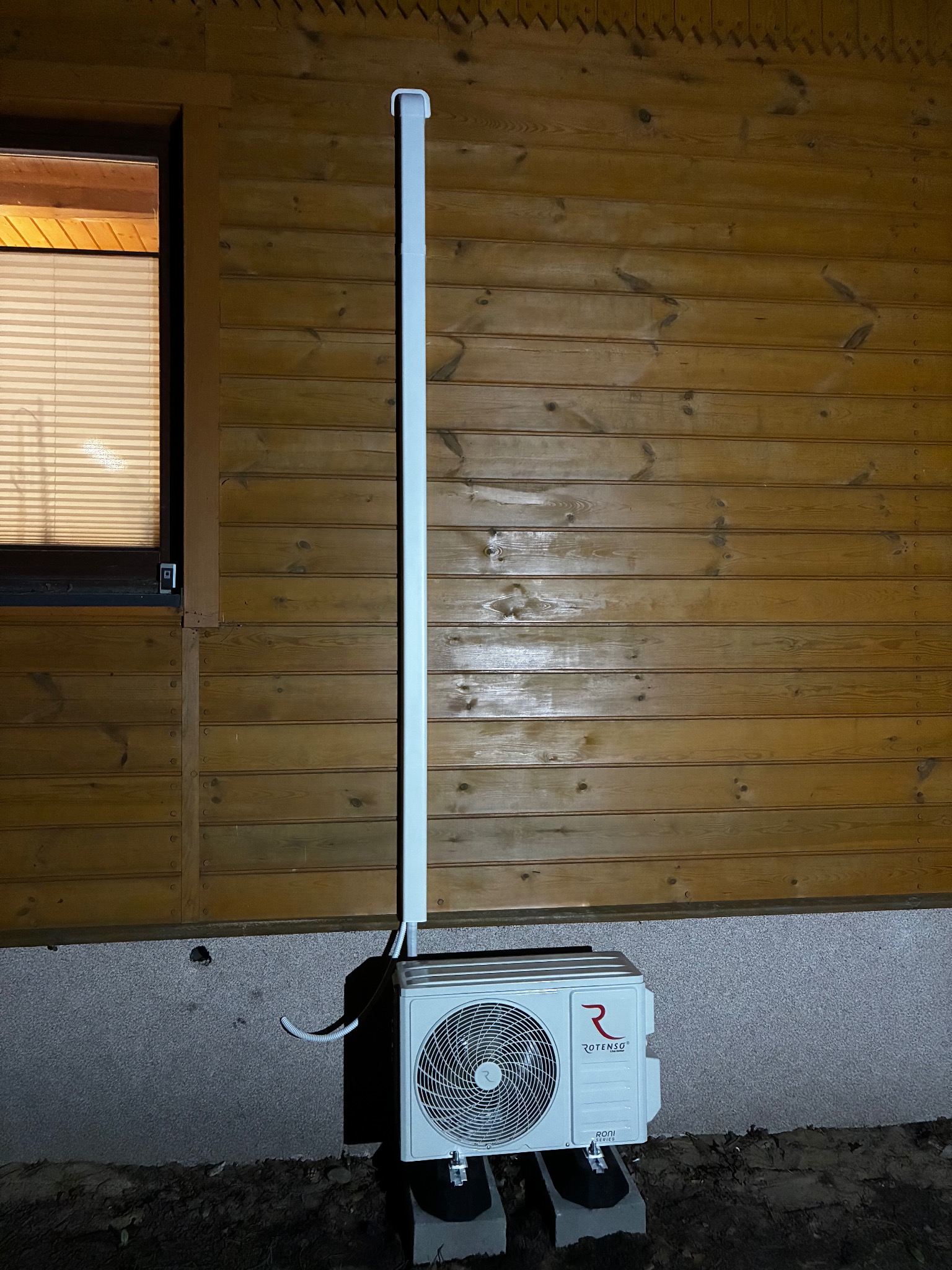 Zamontowana jednostka zewnętrzna klimatyzacji Rotenso przy drewnianej ścianie domu, z widoczną instalacją odprowadzającą skropliny. Ujęcie nocne.